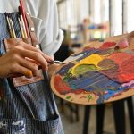 Close-up image of a young creative artist's hands smeared with watercolors holding a spatula mixing color on a palette.
